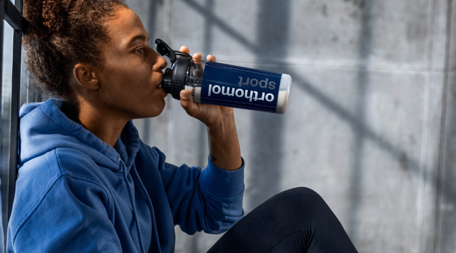 Frau trinkt aus einer Flasche mit Orthomol Sport. Flasche mit weißem Pulver.
